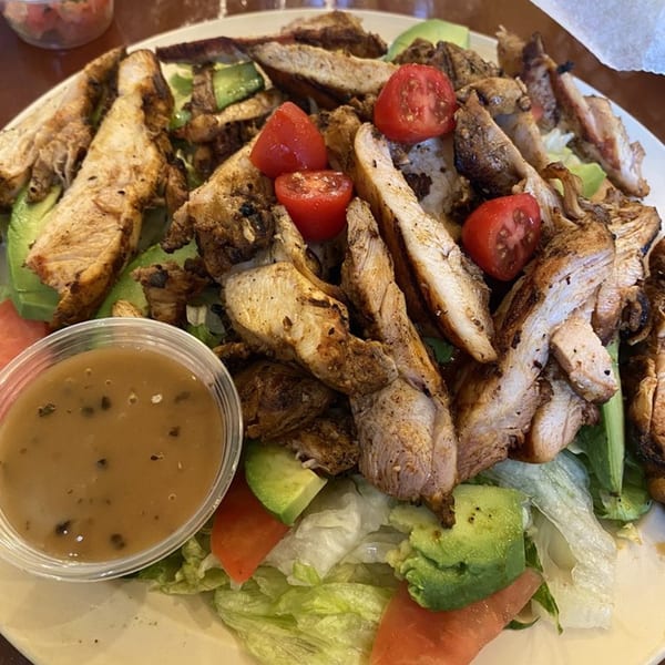 Grilled chicken salad with lettuce, avocado, tomatoes, and a side of dressing.
