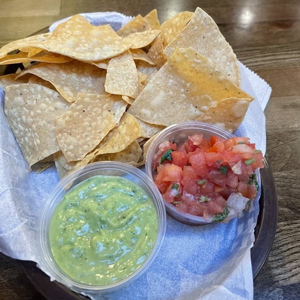 A basket of La Corneta Taqueria tortilla chips with sides of guacamole and pico de gallo.
